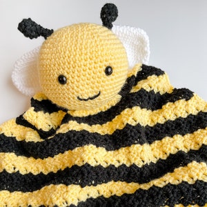 May include: A crocheted bee-shaped baby blanket with yellow and black stripes. The bee has a white face and black eyes.