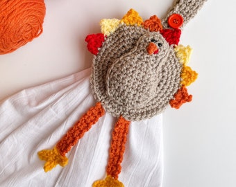 Pode incluir: Um porta-toalhas de peru feito à mão em crochê, com corpo bege, pernas laranja e um botão vermelho. O peru tem um corpo redondo com uma cabeça pequena e uma cauda colorida. O porta-toalhas está preso a uma toalha branca, com lã vermelha, laranja e amarela no fundo.