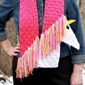 Puede incluir: Una bufanda de crochet rosa y blanca con un diseño de cabeza de unicornio. El unicornio tiene un cuerpo blanco, un cuerno amarillo y una crin rosa, amarilla y verde. La bufanda tiene flecos en los extremos.