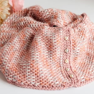 Pode incluir: Uma echarpe de malha rosa e branca com fecho de botão. A echarpe é feita de um fio macio e texturizado e tem um efeito degradê sutil.