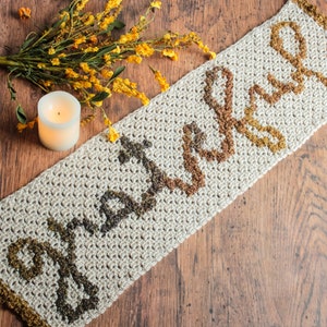 May include: A crocheted beige runner with a brown, orange, and yellow design. The runner has a scalloped edge and is made with a textured stitch.