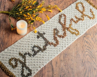 Pode incluir: Um corredor de mesa de crochê bege com um design marrom, laranja e amarelo. O corredor de mesa tem uma borda festonada e é feito com um ponto texturizado.