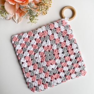 Puede incluir: Un cuadrado de ganchillo rosa, gris y blanco con un anillo de madera adjunto. El cuadrado está hecho con un patrón de punto de abuela.