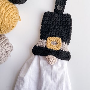 Pode incluir: Um porta-toalhas de croch&ecirc; em forma de gnomo preto e castanho com uma toalha branca pendurada nele. O gnomo tem um chap&eacute;u preto com uma faixa castanha e um nariz de pompom castanho.
