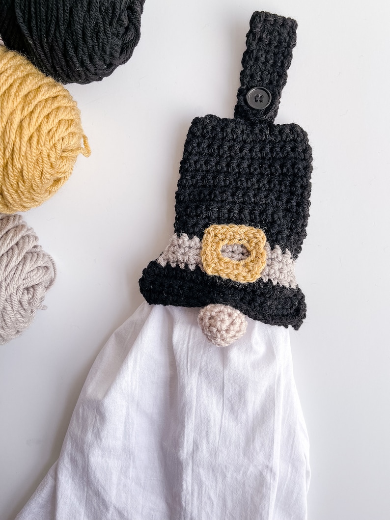 Pode incluir: Um porta-toalhas de croch&ecirc; em forma de gnomo preto e castanho com uma toalha branca pendurada nele. O gnomo tem um chap&eacute;u preto com uma faixa castanha e um nariz de pompom castanho.