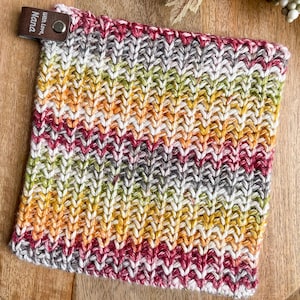 May include: A square, hand-knitted item with a textured pattern in a gradient of colours including red, yellow, green, white, and grey. A small leather tag with the text "With love, Nana" is attached. Likely a washcloth or dishcloth.