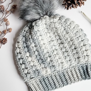 May include: A crocheted beanie in shades of gray and white, featuring a textured puff stitch pattern and a ribbed brim. The hat is topped with a large, fluffy gray faux fur pom-pom. The design incorporates a variegated yarn color scheme.