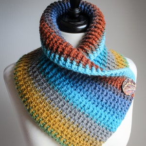 May include: A colorful crocheted cowl with a brown, blue, gray, yellow, and green color scheme. The cowl has a decorative button with a paw print design.