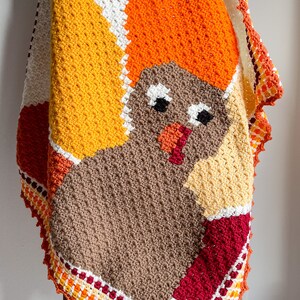 May include: A crocheted baby blanket featuring a colorful turkey design. The blanket is made with brown, orange, yellow, red, and white yarn. The turkey has a brown body, orange head, and yellow feathers. The blanket has a scalloped edge.