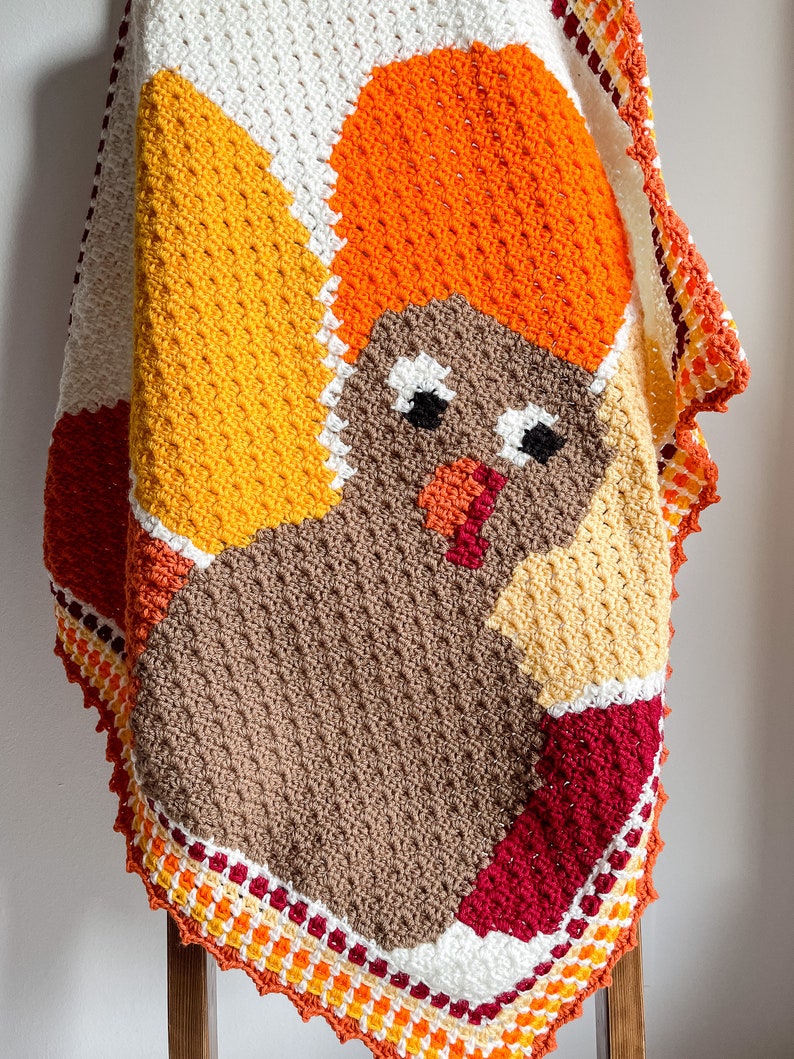 May include: A crocheted baby blanket featuring a colorful turkey design. The blanket is made with brown, orange, yellow, red, and white yarn. The turkey has a brown body, orange head, and yellow feathers. The blanket has a scalloped edge.