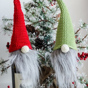 Op de afbeelding: Twee gehaakte gnomen, een rode en een groene, worden gebruikt als flessenstoppers. De gnomen hebben witte pompoenen voor neuzen en grijze bontbaarden. De gnomen staan voor een kleine kunstkerstboom.