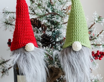 May include: Two crocheted gnomes, one red and one green, are used as bottle stoppers. The gnomes have white pom-poms for noses and gray fur beards. The gnomes are placed in front of a small artificial Christmas tree.