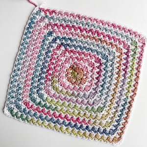 Op de afbeelding: Een vierkant gehaakt granny square met een veelkleurig midden en een witte rand. Het midden is een verloop van roze, oranje, geel en groen.