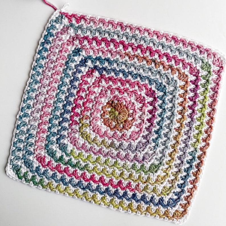 Op de afbeelding: Een vierkant gehaakt granny square met een veelkleurig midden en een witte rand. Het midden is een verloop van roze, oranje, geel en groen.