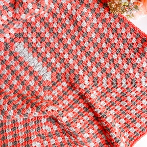 Op de afbeelding: Een handgemaakte gehaakte deken in een granny square patroon. De deken heeft een herhalend patroon van koraal, grijze en witte vierkanten. De deken is opgevouwen, wat de textuur- en kleurvariaties laat zien. Het algemene thema is gezellig en handgemaakt.