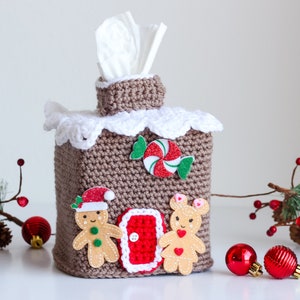 Op de afbeelding: Gehaakte gingerbread house tissuebox cover met een wit gehaakt dak, een rode en witte pepermuntstok, gingerbread poppetjes en een rode deur.