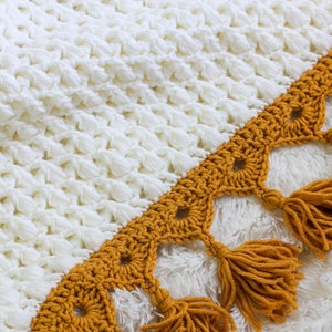 May include: A white crocheted blanket with a mustard yellow crocheted border and tassels.