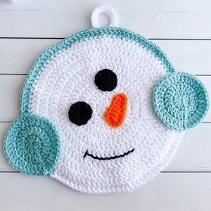 Puede incluir: Un adorno de muñeco de nieve de crochet blanco con una boca negra, ojos negros y una nariz naranja. El muñeco de nieve tiene dos orejeras de crochet de color turquesa.