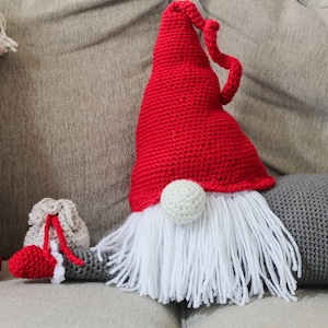 May include: A red and white crocheted gnome pillow with a long white beard. The gnome is sitting on a gray couch with a red pillow with white snowflakes behind it.