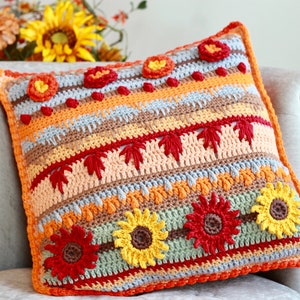 May include: A crocheted pillow with a fall theme. The pillow is decorated with sunflowers, leaves, and other fall-themed motifs in shades of orange, yellow, red, brown, and green.
