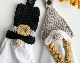 Pode incluir: Dois toppers de toalha de gnomo de crochê, um em preto e marrom com um cinto e outro em cinza e branco com cabelo trançado amarelo.