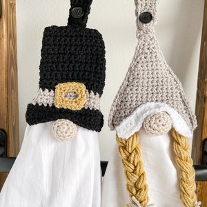 Pode incluir: Dois toppers de toalha de gnomo de croch&ecirc; pendurados em um rack de metal preto. Um gnomo tem um chap&eacute;u preto com uma fivela dourada e uma barba branca. O outro gnomo tem um chap&eacute;u cinza com uma barba branca e um longo cabelo tran&ccedil;ado amarelo.