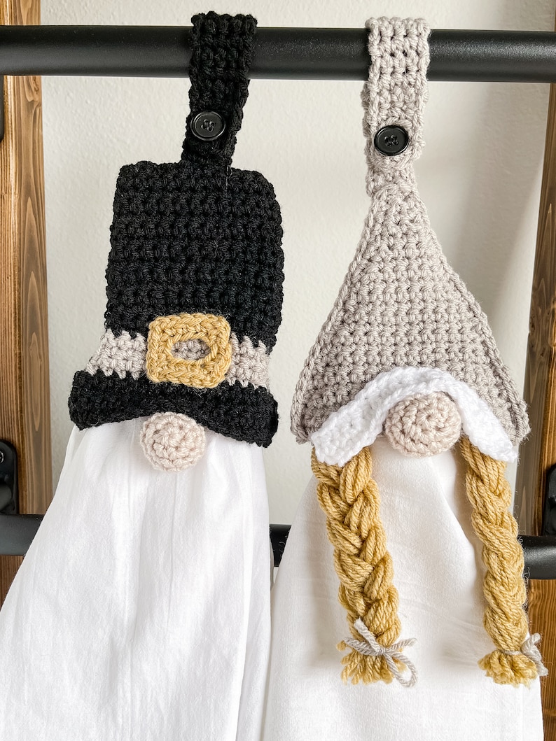 Pode incluir: Dois toppers de toalha de gnomo de croch&ecirc; pendurados em um rack de metal preto. Um gnomo tem um chap&eacute;u preto com uma fivela dourada e uma barba branca. O outro gnomo tem um chap&eacute;u cinza com uma barba branca e um longo cabelo tran&ccedil;ado amarelo.