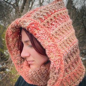 May include: A handmade crochet hood in shades of peach, cream, and brown. The hood is designed with a ribbed texture and a pointed top, offering warmth and style. The hood is a cozy accessory for cold weather.