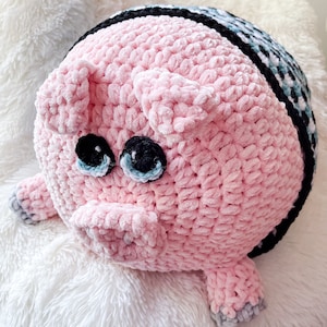 May include: A handmade, plush pink pig-shaped pillow. The pig has black and blue eyes, a pink snout, and small ears. The pillow is made of soft, textured yarn and has gray hooves. The top of the pig has a black and blue patterned design.