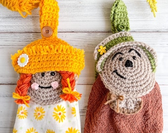 May include: Two crocheted towel toppers, one with a brown face and a brown hat with a green brim, and the other with a brown face and a yellow hat with a white flower. Both toppers have orange hair and are hanging on a white background.