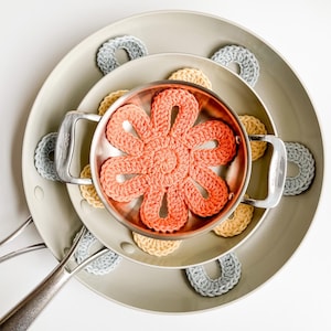 May include: A collection of handmade crochet items, including a large coral flower-shaped trivet inside a copper-colored pan, surrounded by gray, yellow, and gray circular coasters. The items are arranged on top of gray pans.