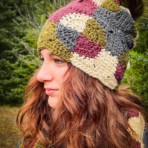 May include: A handmade crochet hat with a matching scarf. The hat features a patchwork design in shades of olive green, gray, cream, and burgundy. A fluffy, olive green pom-pom tops the hat. The scarf is visible at the bottom of the image.