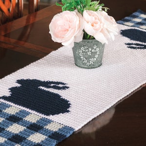 Op de afbeelding: Een witte gehaakte tafelloper met een blauw-wit geruit patroon. Twee blauwe konijnen silhouetten zijn geborduurd op de tafelloper. Een boeket roze bloemen in een metalen pot staat op de tafelloper.