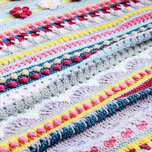 Puede incluir: Una manta de ganchillo colorida con un patrón a rayas y una variedad de texturas, incluyendo flores, pompones y corazones.