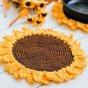 May include: A crocheted sunflower pot holder with a brown center and yellow petals. The pot holder is on a white wooden surface.