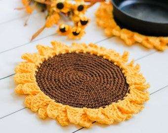 May include: A crocheted sunflower pot holder with a brown center and yellow petals. The pot holder is on a white wooden surface.
