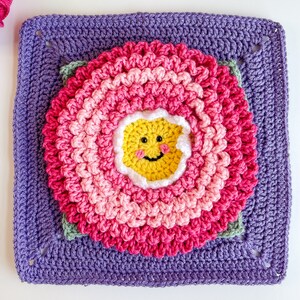 May include: A purple crochet square with a pink and white crocheted flower in the center. The flower has a yellow smiley face in the middle.