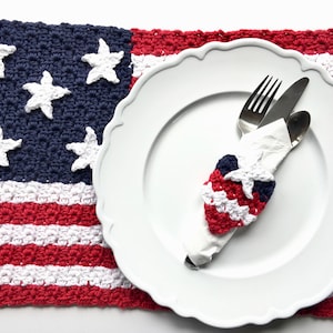 May include: A white plate with silverware and a napkin ring sits on a crocheted placemat. The placemat is designed to resemble the American flag, with red and white stripes and white stars on a blue field. The napkin ring is red, white, and blue.