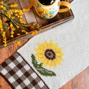 May include: Cream-colored crochet table runner featuring a large yellow sunflower with a brown center and green leaf. The runner has a brown and white checkered pattern on one end. A mug with a sunflower design and a tray with yellow flowers are in the background.