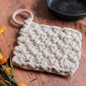 May include: A white crocheted pot holder with a wooden ring handle. The pot holder is on a wooden surface with a cast iron skillet in the background.