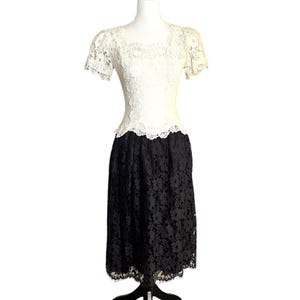 May include: A two-tone dress featuring a white lace bodice with short sleeves and a square neckline, paired with a black lace skirt that falls to a midi length. The bodice and skirt are joined at the waist with a scalloped lace edge detail.