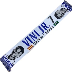 May include: White and blue scarf with a photo of a soccer player and the text "VINI JR. 7 MADRID & BRASIL" printed on it.
