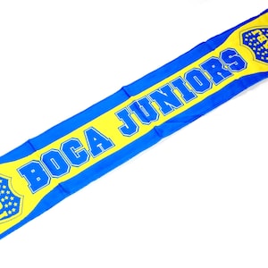 Può includere: Una sciarpa blu e gialla con le parole "Boca Juniors" e un logo con le lettere "CABJ" in uno scudo.