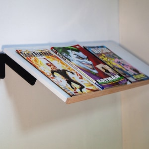 May include: A white shelf with a wood edge, mounted to a white wall with a black metal bracket. Three comic books are on the shelf, including "Black Panther", "Batman The Killing Joke", and "The Joker The Man Who Laughs".