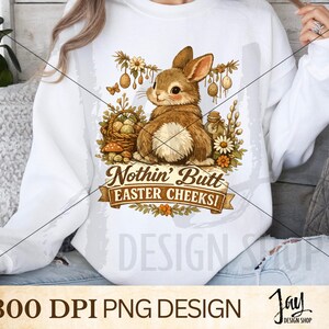 Könnte beinhalten: Weißes Sweatshirt mit einer braunen Hasenillustration und dem Text "Nothin' Butt Easter Cheeks!". Das Design umfasst einen Korb mit Eiern, Blumen und dekorativen Elementen, ideal für Ostern.