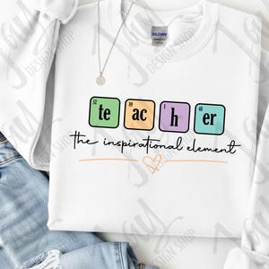 Può includere: Una felpa bianca a girocollo con un design grafico in cui la parola teacher è scritta all'interno di riquadri colorati che ricordano la tavola periodica. Sotto il testo, la frase the inspirational element è scritta in corsivo nero con un piccolo cuore arancione.