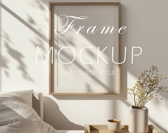 May include: A wooden frame mockup with a white mat and a light brown wood frame. The frame is hanging on a white wall with a white couch and a tray with a cup and a vase of dried flowers in front of it.