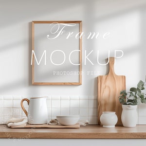 Puede incluir: Una cocina blanca con una maqueta de marco de madera, una tabla de cortar de madera, una jarra blanca, dos tazones blancos y un jarrón blanco con hojas verdes. El texto "Frame Mockup Photoshop File" está escrito en el marco.
