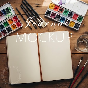 May include: An open journal with a brown bookmark, surrounded by watercolor paint sets and paintbrushes on a wooden surface. The text "Journal Mockup" is overlaid on the image.