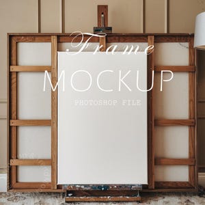 Puede incluir: Una maqueta de marco de lienzo blanco en un caballete, con un marco de madera y un fondo marrón claro. El texto "Frame Mockup" y "Photoshop File" está impreso en el lienzo.